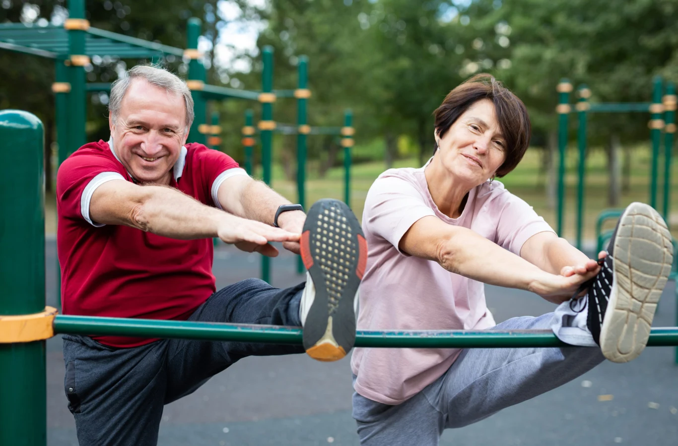 Ćwiczenia dla seniorów w polskich parkach – bezpiecznie i skutecznie Dwoje starszych ludzi w sportowych ubraniach wykonuje rozciąganie nóg na drążku w parku, oboje uśmiechnięci i skoncentrowani na ćwiczeniach.