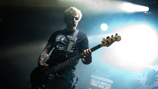 muzyk grający na gitarze basowej na scenie, dynamiczne oświetlenie skierowane zza jego pleców, widoczny wzmacniacz i sprzęt muzyczny w tle