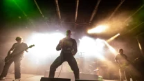 Zespół rockowy grający koncert na scenie, centralnie wokalista w dynamicznej pozie, po bokach gitarzyści i basista, za nimi intensywne światła sceniczne i dym tworzący energetyczną atmosferę.