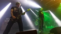 dwóch członków zespołu rockowego występuje na scenie w dynamicznym oświetleniu, jeden gra na gitarze basowej, drugi trzyma mikrofon, w tle perkusja oraz dym i kolorowe światła tworzące koncertowy klimat