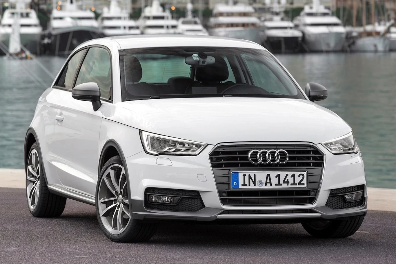 Audi A1 z rocznika 2015 biały samochód marki Audi stoi zaparkowany na tle przystani z luksusowymi jachtami, jego sportowy wygląd podkreślają charakterystyczne reflektory LED oraz srebrne felgi