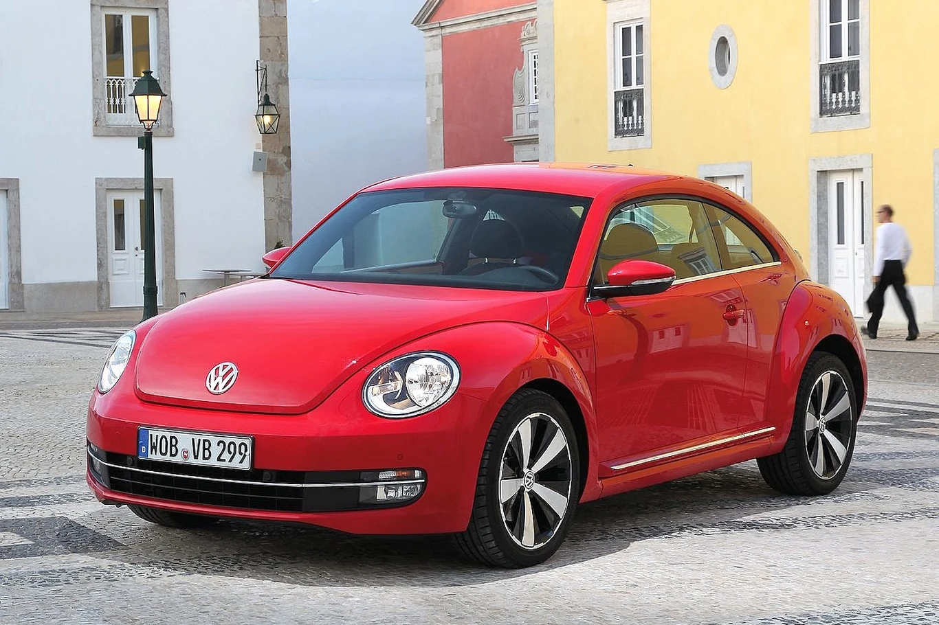 Volkswagen Beetle rocznik 2012 czerwony Volkswagen Beetle stoi na brukowanym placu, w tle kolorowe elewacje budynków oraz przechodzień