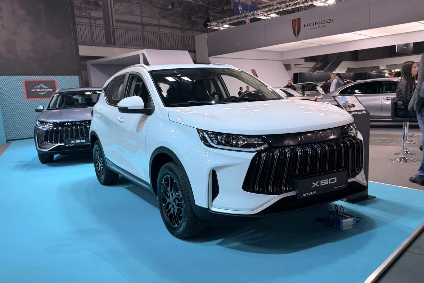Jetour X50 to nowy najtańszy model marki Jetour X50 biały SUV z czarnym grillem i felgami na niebieskiej platformie, obok ciemny model tej samej marki