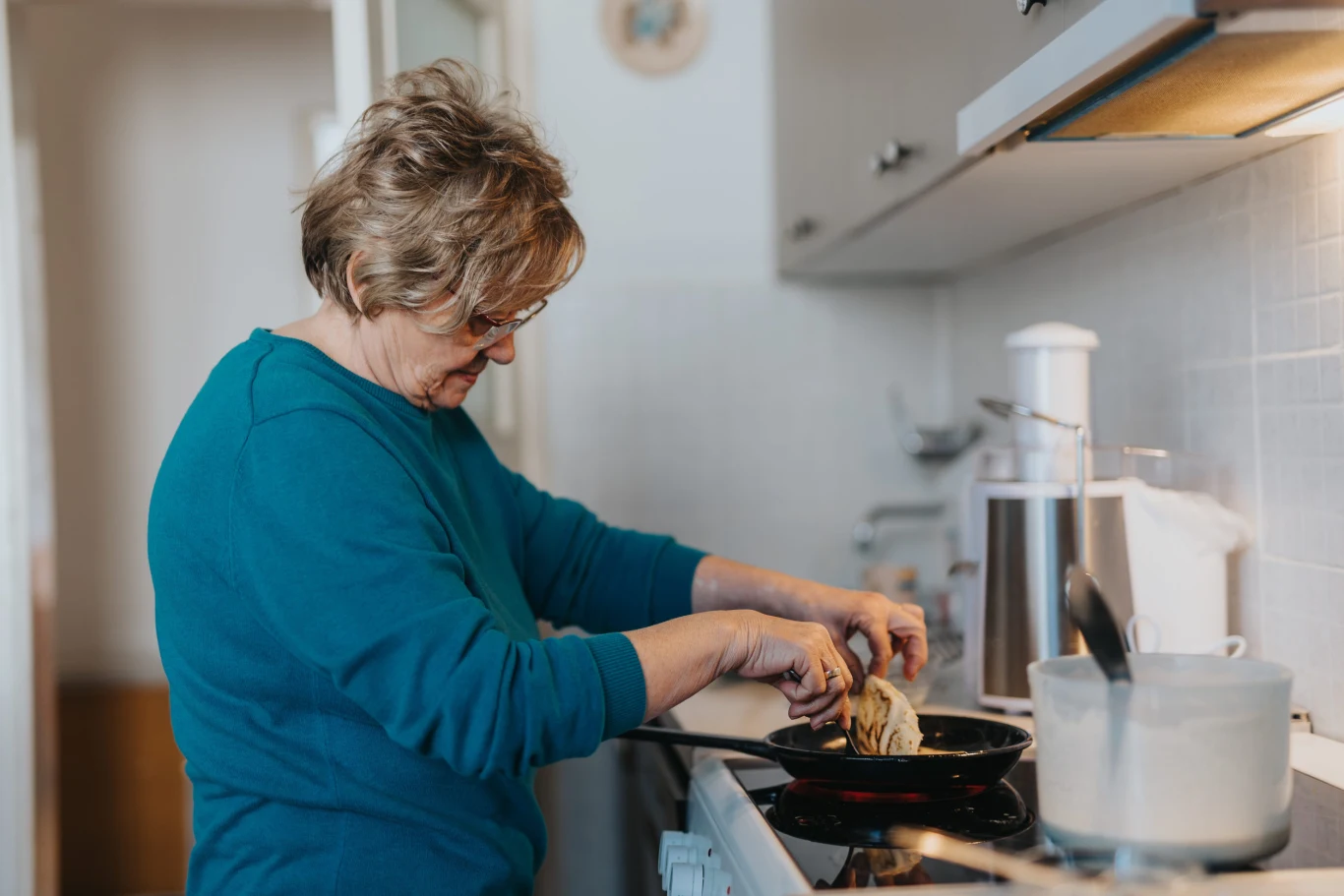 starsza kobieta w okularach smaży jedzenie na patelni w kuchni, skupiając się na przygotowaniu posiłku