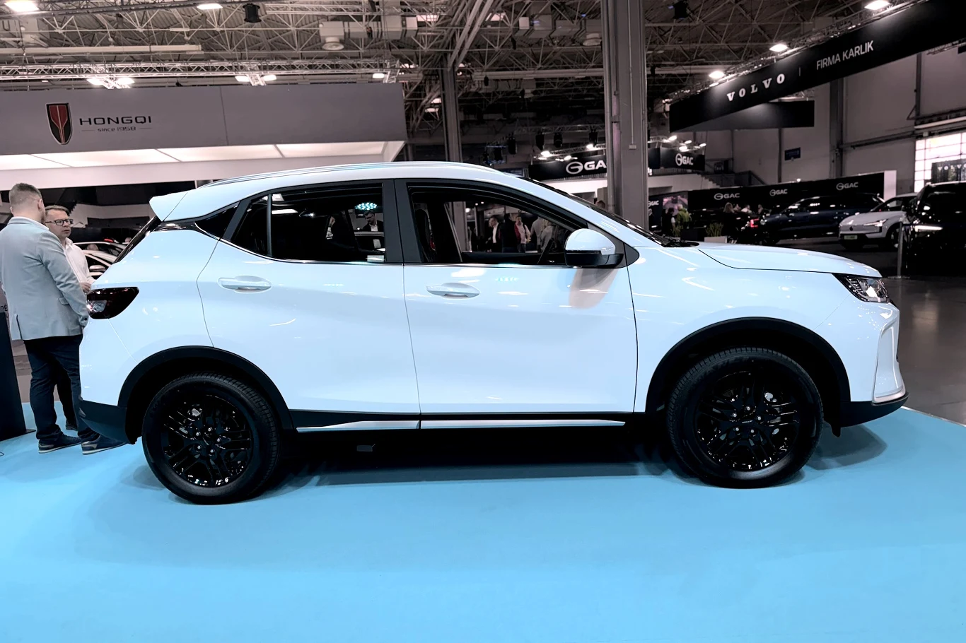 Jetour X50 biały nowoczesny SUV z czarnymi felgami prezentowany na targach motoryzacyjnych, w tle stoiska z innymi samochodami oraz logotypami marek motoryzacyjnych