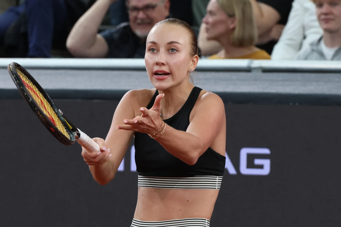 tenisistka w czarnym stroju sportowym gestykuluje dłonią i trzyma rakietę, wyrażając silne emocje podczas meczu na tle skupionej publiczności