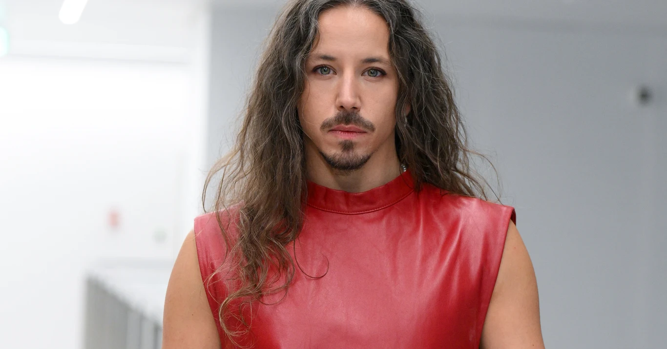 Michał Szpak Michał Szpak w czerwonym skórzanym bezrękawniku, z długimi kręconymi włosami, na tle jasnego wnętrza