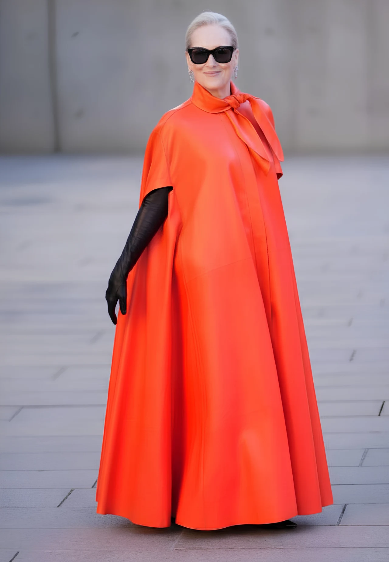 kobieta ubrana w długą, pomarańczową pelerynę z kokardą pod szyją, czarne rękawiczki i duże ciemne okulary przeciwsłoneczne, stoi na jasnej, gładkiej powierzchni