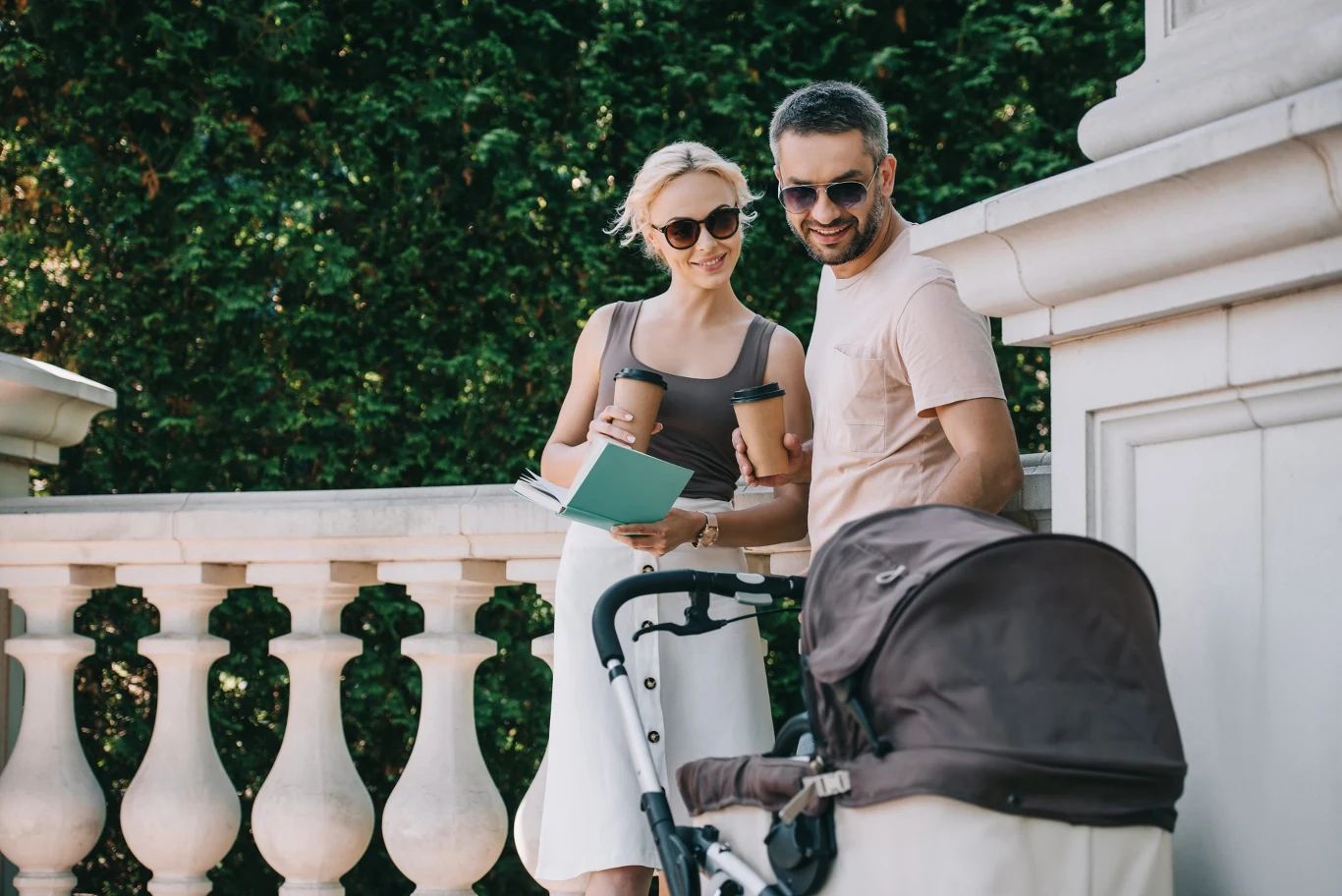 Krakowianie nadają swoim dzieciom stare, klasyczne imiona, Młoda para z wózkiem, kubkami kawy i dokumentami spaceruje obok białej balustrady w Krakowie.