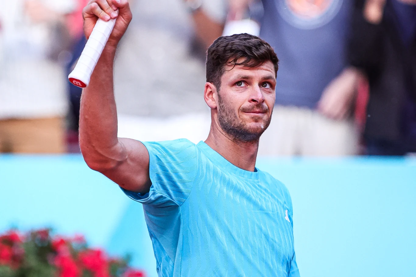 tenisista w niebieskiej koszulce trzyma rakietę uniesioną w górę, patrzy w stronę publiczności z poważnym wyrazem twarzy, w tle rozmazane sylwetki kibiców i czerwone kwiaty