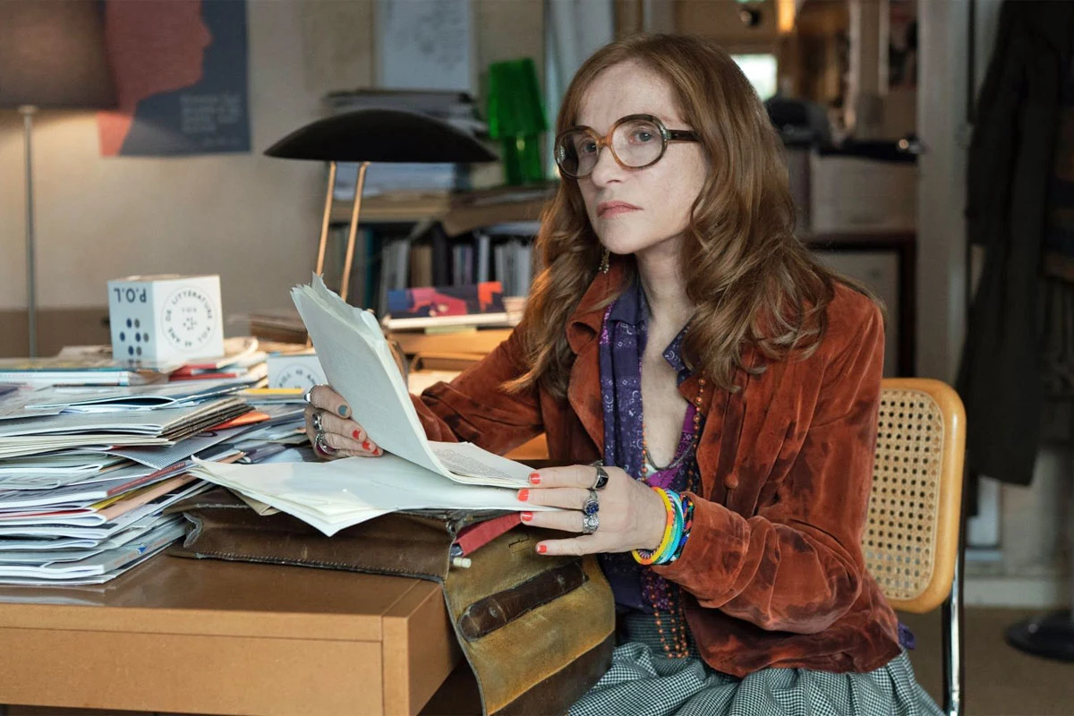 Isabelle Huppert w scenie z filmu "Historie równoległe" Isabelle Huppert przy biurku ze stosami dokumentów i czasopism w filmie "Historie równoległe".