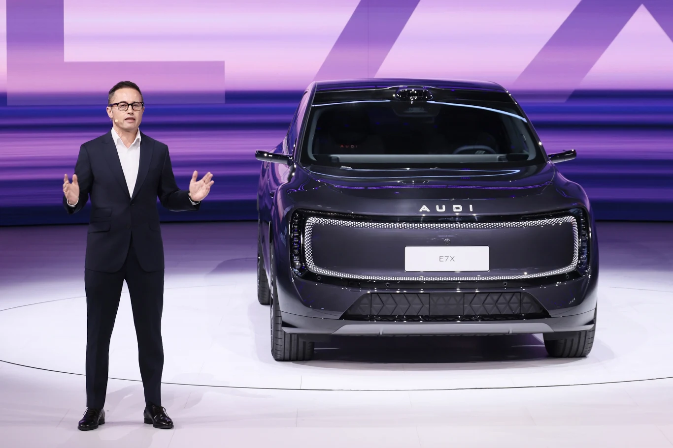 Audi E7X zadebiutowało na salonie w Pekinie Mężczyzna w garniturze prezentuje Audi E7X z dużym grillem na tle fioletowo-różowego podświetlenia w Pekinie.