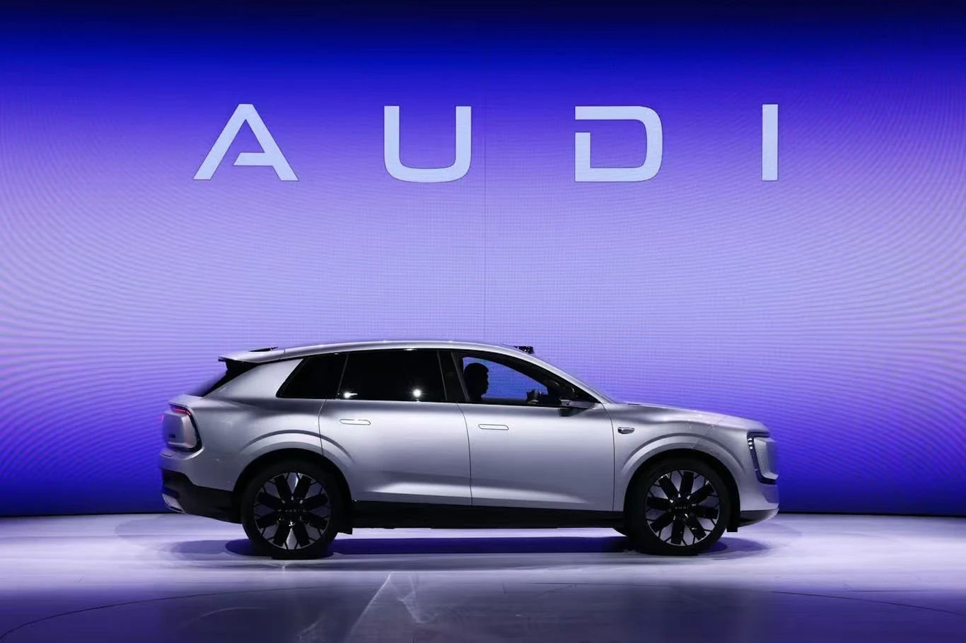 Audi E7X srebrny samochód marki Audi ustawiony bokiem na tle dużego, podświetlonego napisu AUDI na niebieskim tle