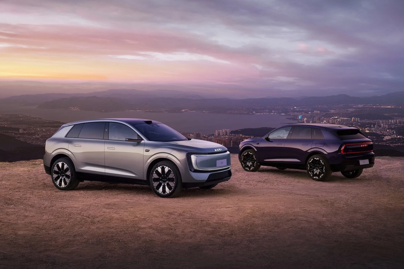 Audi E7X Dwa nowoczesne SUV-y zaparkowane na szczycie wzgórza z panoramicznym widokiem na miasto o zmierzchu, podkreślone stylowym oświetleniem i wyrazistym designem pojazdów.