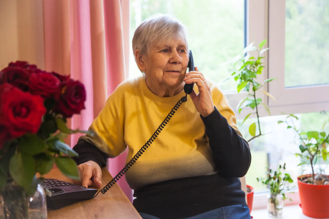 Starsza kobieta w żółto-czarnym swetrze siedzi przy stole i rozmawia przez telefon stacjonarny, w tle widoczne są doniczki z roślinami na parapecie oraz bukiet czerwonych róż po lewej stronie kadru.
