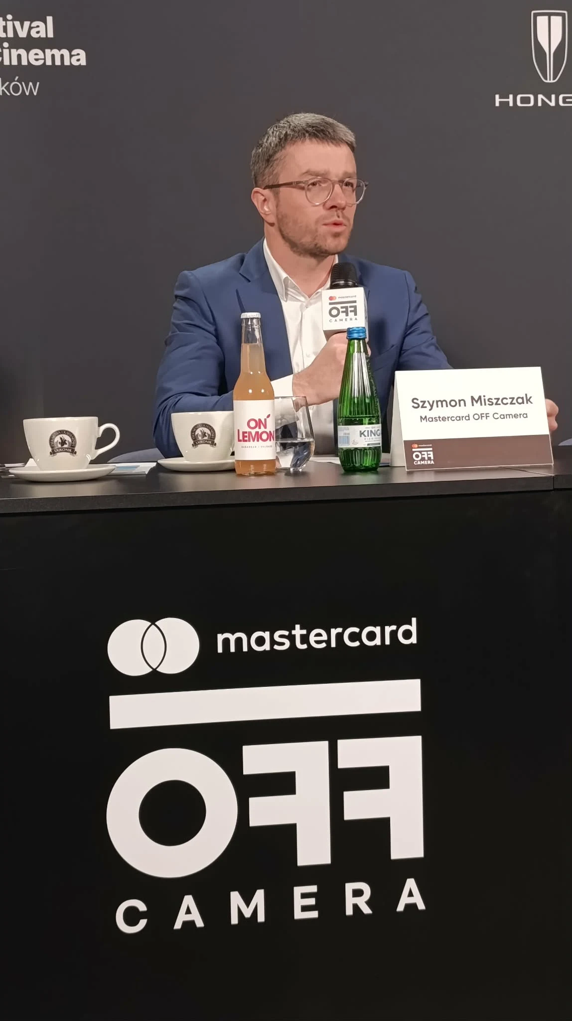 Szymon Miszczak - dyrektor festiwalu Mastercard OFF CAMERA mężczyzna w granatowej marynarce i okularach siedzi za stołem konferencyjnym, przed nim są mikrofon, butelka wody, napój, filiżanki i tabliczka z imieniem oraz nazwiskiem, tło z logotypami Mastercard OFF Camera i innych sponsorów