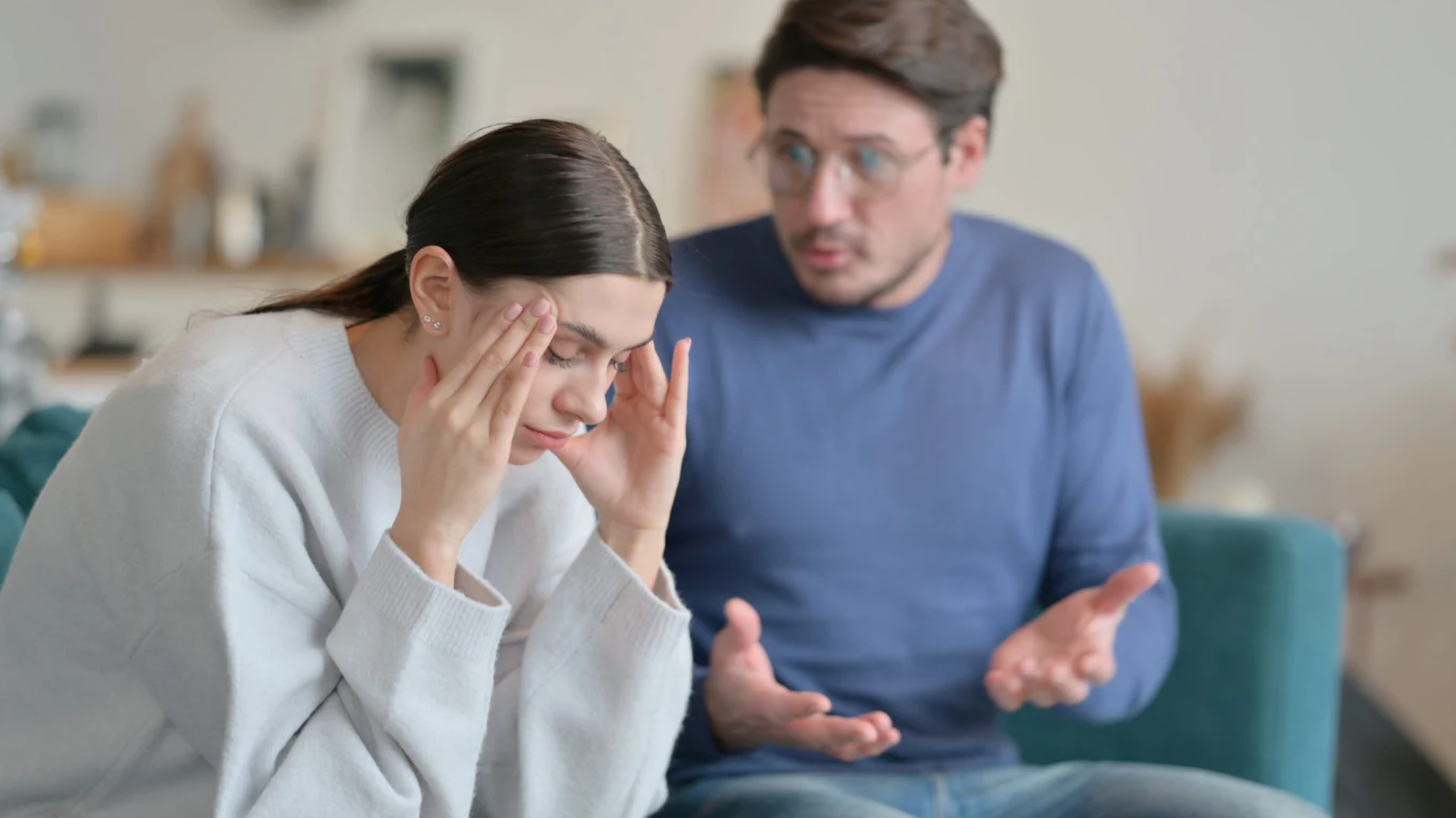 kobieta siedzi z głową opartą o ręce, wyraźnie zestresowana lub zmartwiona, podczas gdy mężczyzna obok niej gestykuluje i wygląda na zdezorientowanego lub zatroskanego; atmosfera sugeruje napiętą rozmowę lub konflikt