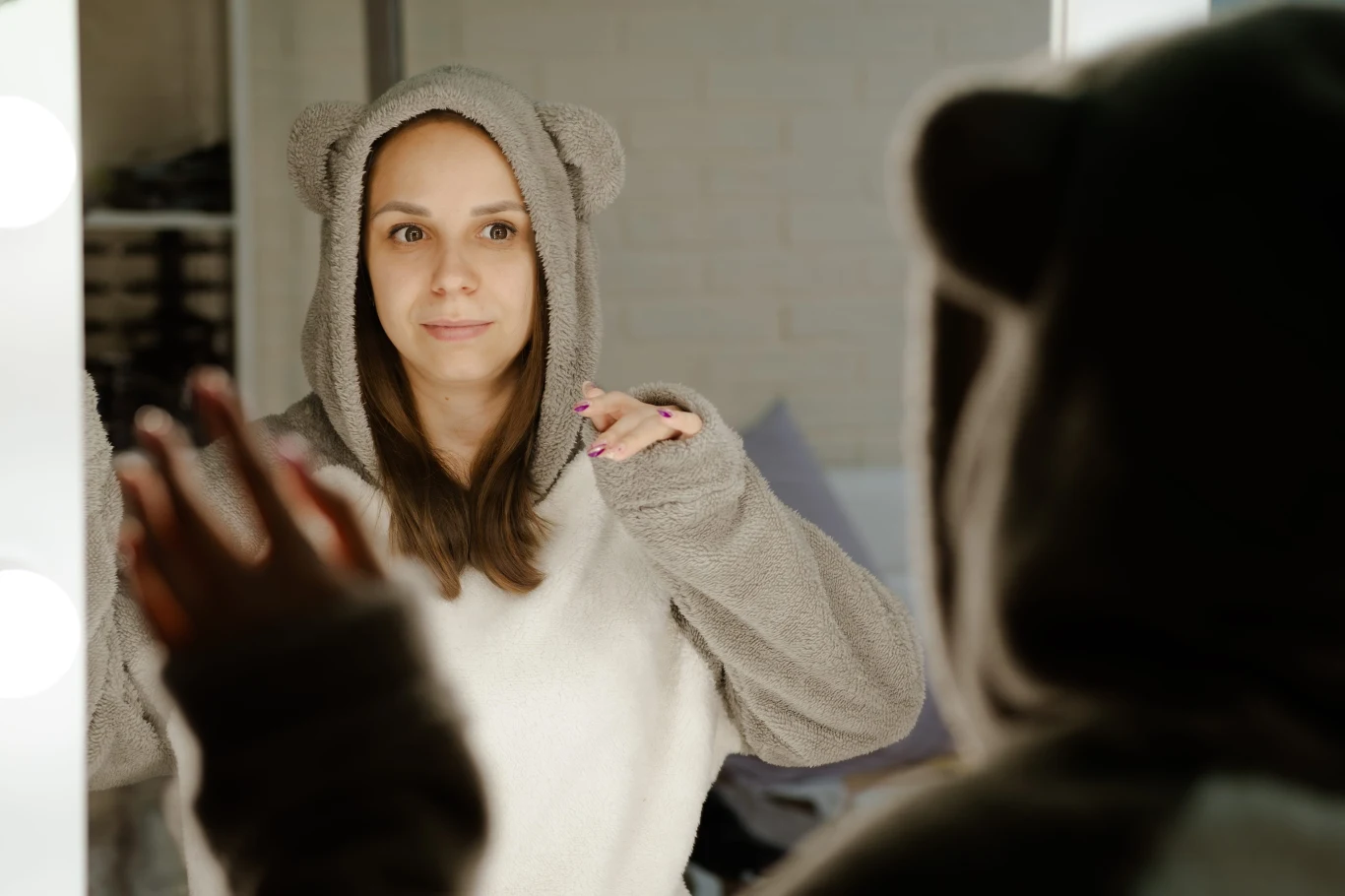 Luźne, miękkie, często pluszowe bluzy czy kombinezony chętnie są wybierane jako "podomki" przez panie kobieta w szarym kombinezonie z kapturem z uszami patrzy na swoje odbicie w lustrze, dotykając ręką jego powierzchni