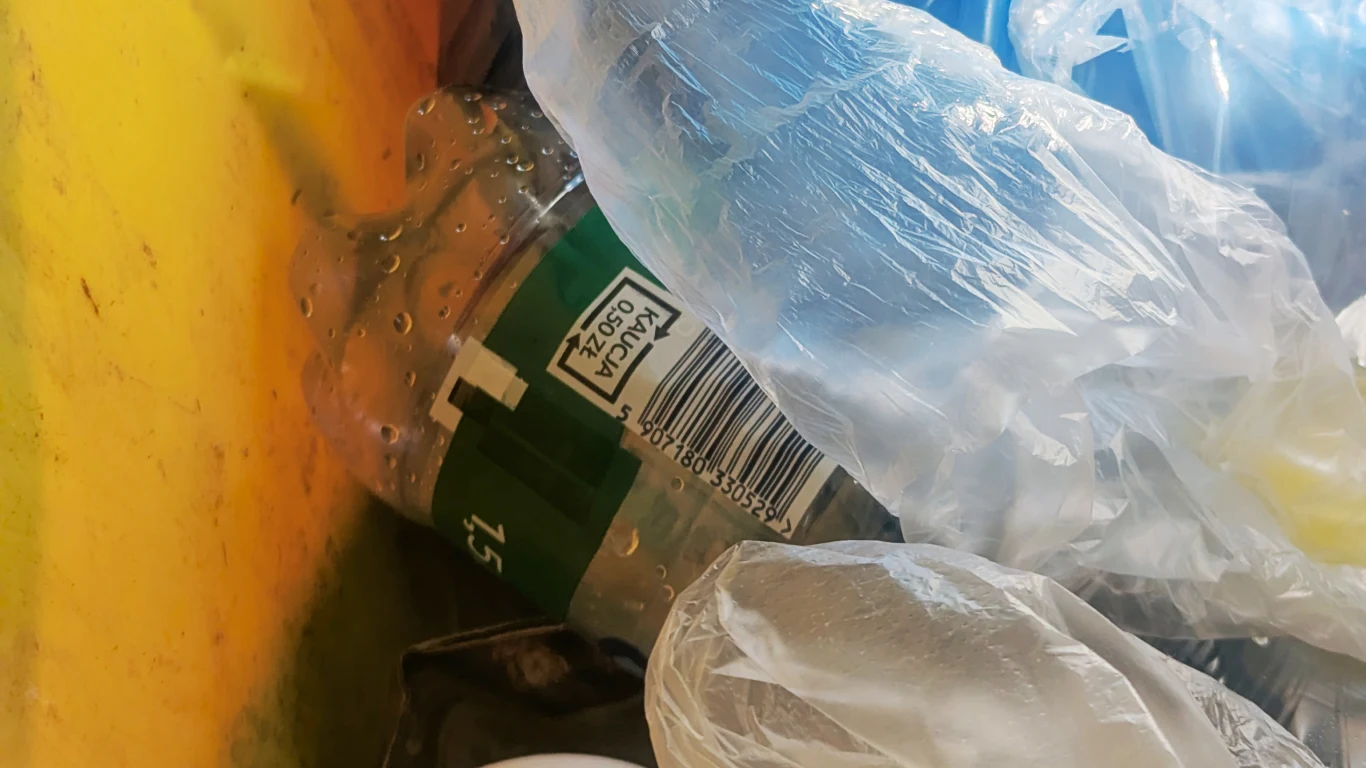 Plastikowa butelka po wodzie mineralnej i kilka przezroczystych foliowych reklamówek znajdują się w żółtym pojemniku na odpady, wskazując na selektywną zbiórkę plastiku.