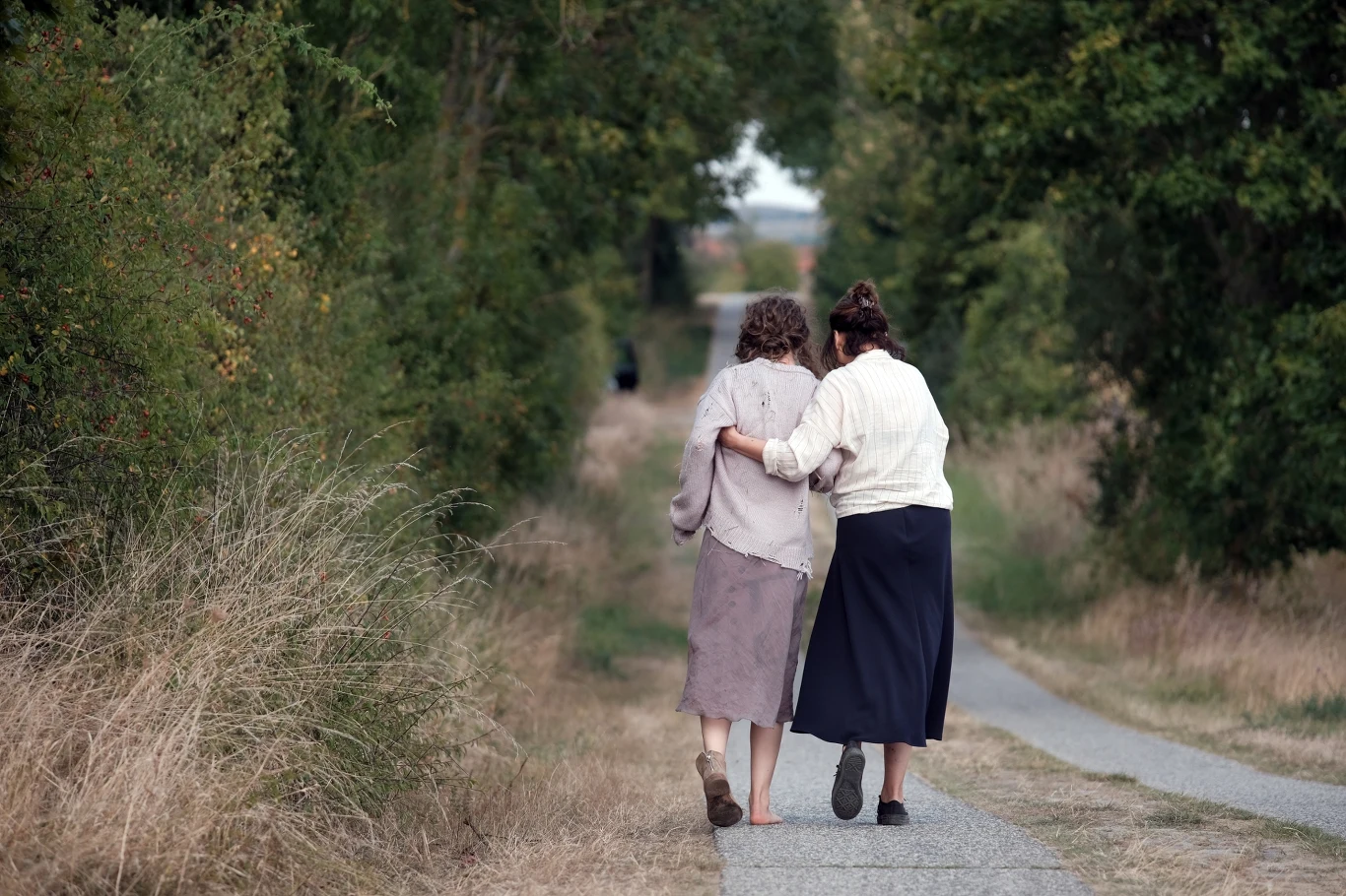 Kadr z filmu "Miroirs III. Barka na oceanie" dwie kobiety idące razem wzdłuż wąskiej, wiejskiej drogi, obejmujące się ramionami, otoczone wysoką trawą i drzewami po obu stronach ścieżki