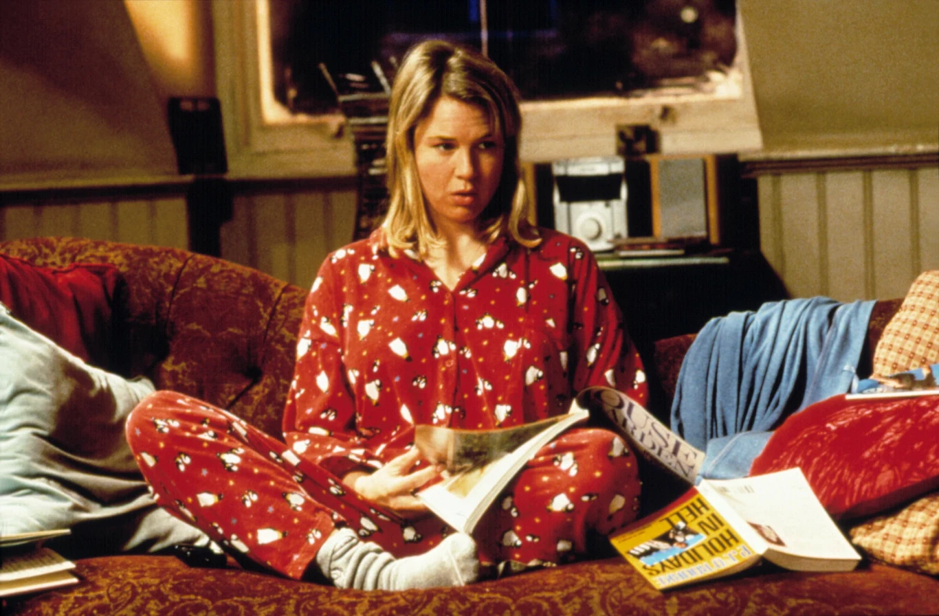 Uwielbiana postać Bridget Jones w "domowej wersji" kobieta w czerwonej piżamie w białe wzory siedzi po turecku na kanapie z rozłożonymi wokół siebie książkami i czasopismami, z wyrazem zamyślenia na twarzy