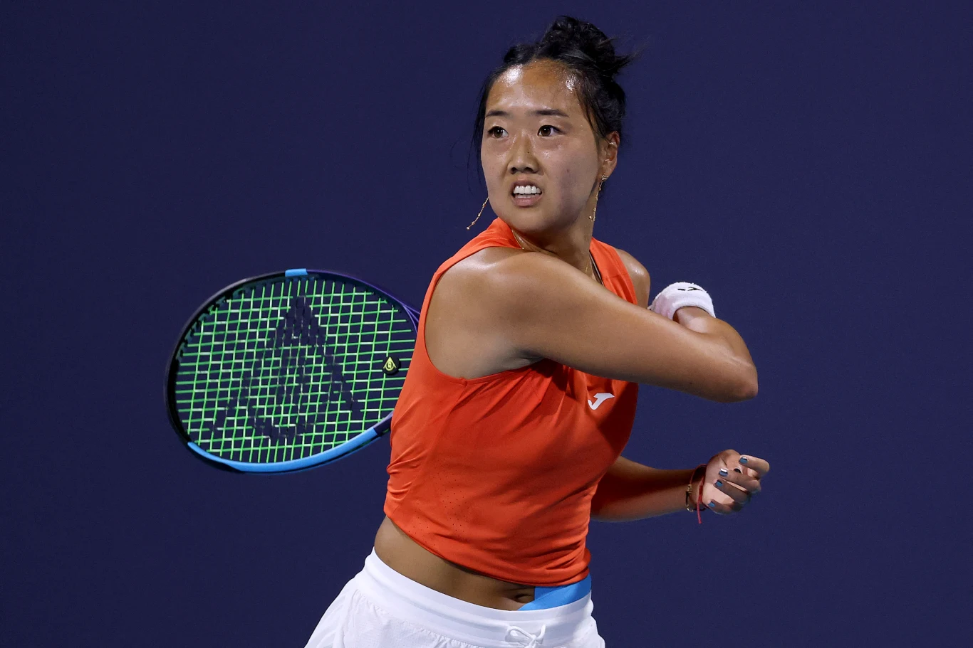 tenisistka w pomarańczowej koszulce i białej spódnicy wykonuje zamach rakietą podczas meczu na korcie, na tle ciemnoniebieskiego tła