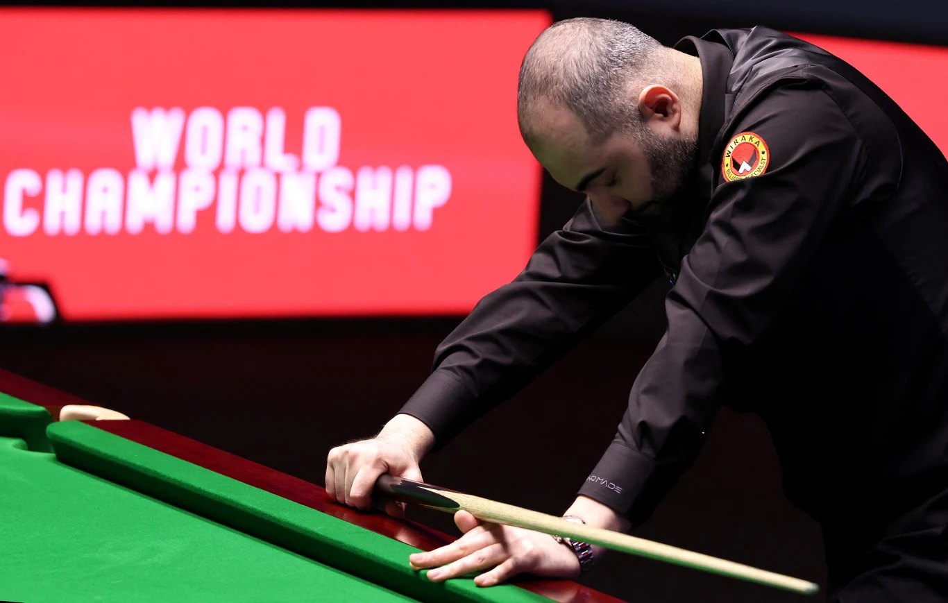 Zawodnik pochylony nad stołem bilardowym w trakcie przygotowania do uderzenia, w tle tablica z napisem „World Championship” oraz sportowa atmosfera podkreślająca rangę wydarzenia.