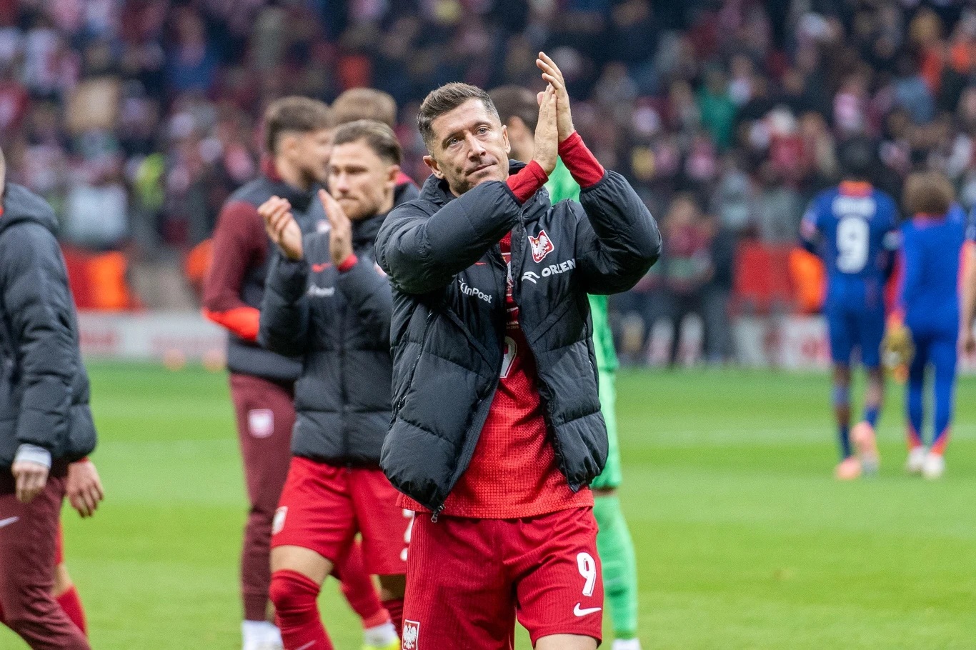 Robert Lewandowski Robert Lewandowski w czerwonym stroju z numerem 9 i czarnej kurtce bije brawo na stadionie wśród piłkarzy i kibiców.