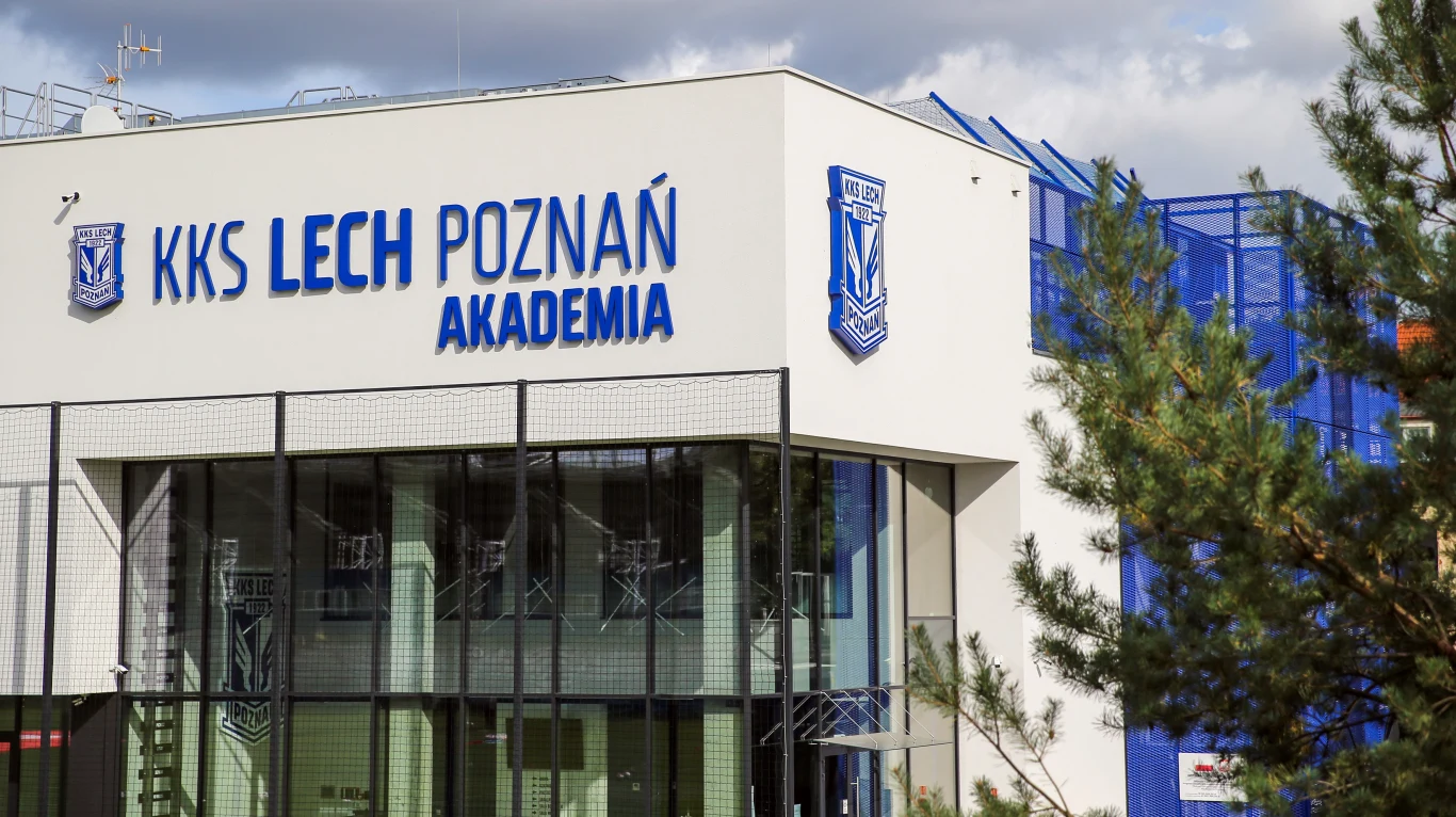 nowoczesny budynek Akademii KKS Lech Poznań z dużym logotypem i napisem na fasadzie, przeszkloną częścią dolną oraz widocznymi elementami infrastruktury sportowej w tle