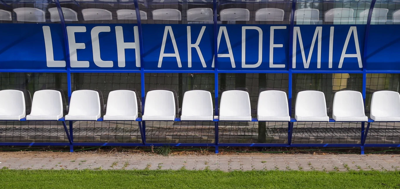 rząd białych plastikowych krzesełek pod niebieskim zadaszeniem z dużym napisem „LECH AKADEMIA” na tle, stojących na brukowanym chodniku przy boisku sportowym