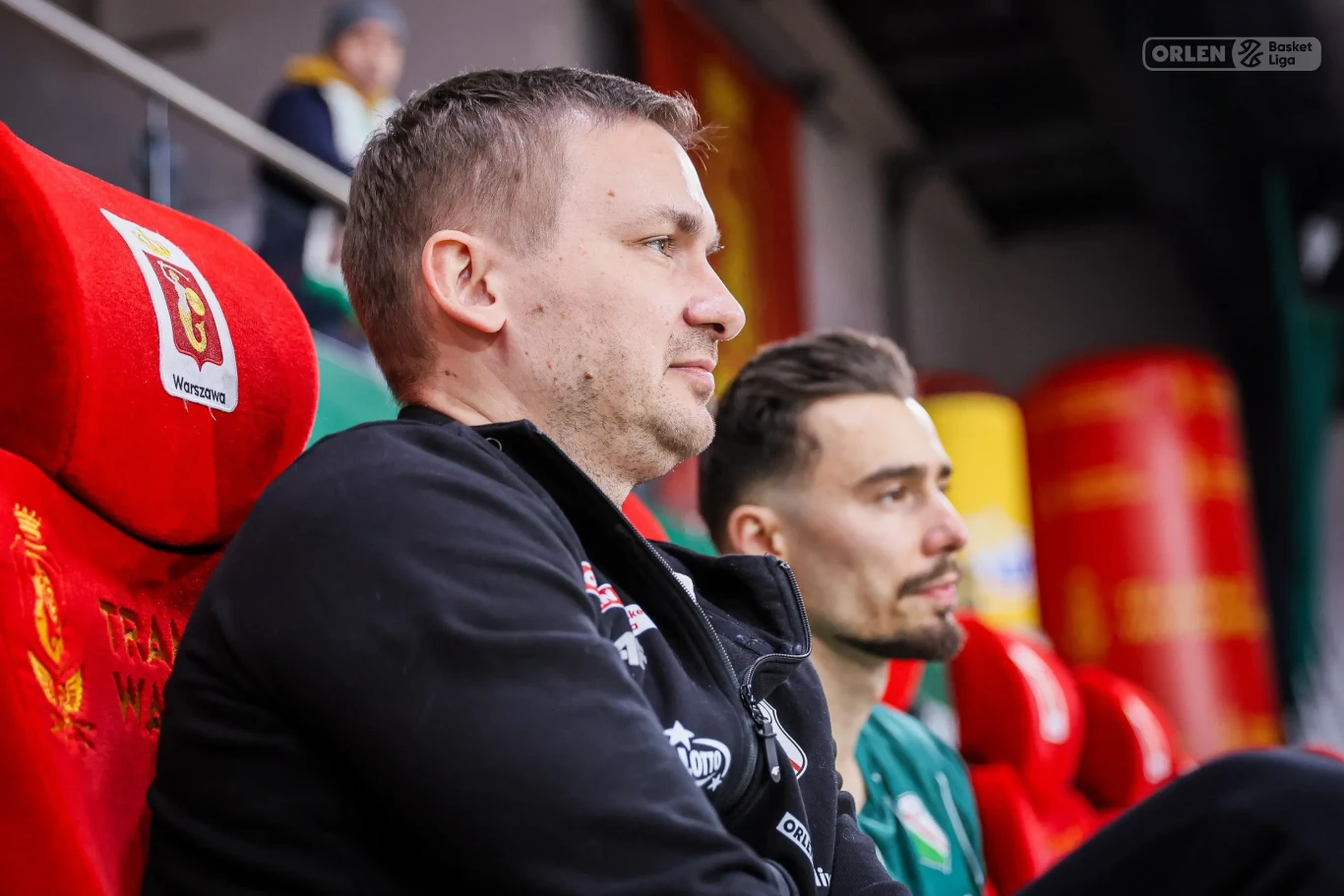 Dwaj mężczyźni siedzą obok siebie na sportowych, czerwonych fotelach z herbami drużyny, koncentrując się na wydarzeniach rozgrywających się przed nimi, w tle widoczne są kolorowe elementy hali sportowej.