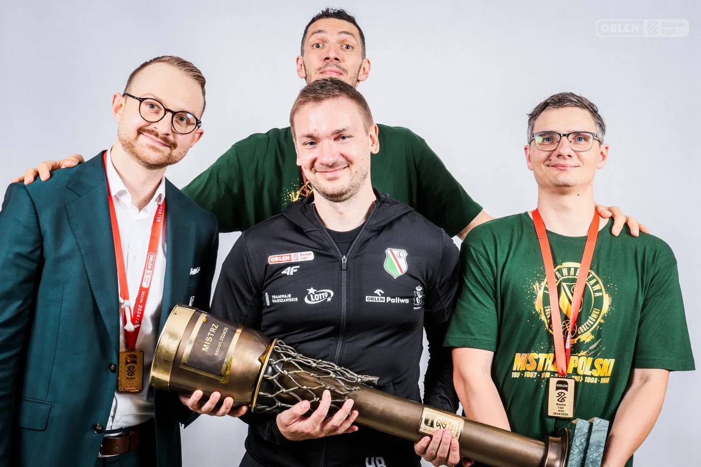 czterech mężczyzn pozuje razem, z czego jeden trzyma dużą sportową statuetkę w kształcie pucharu oplecionego siatką; wszyscy mają na szyjach złote medale, a jeden z nich ubrany jest w oficjalną koszulkę ze sportowym logo