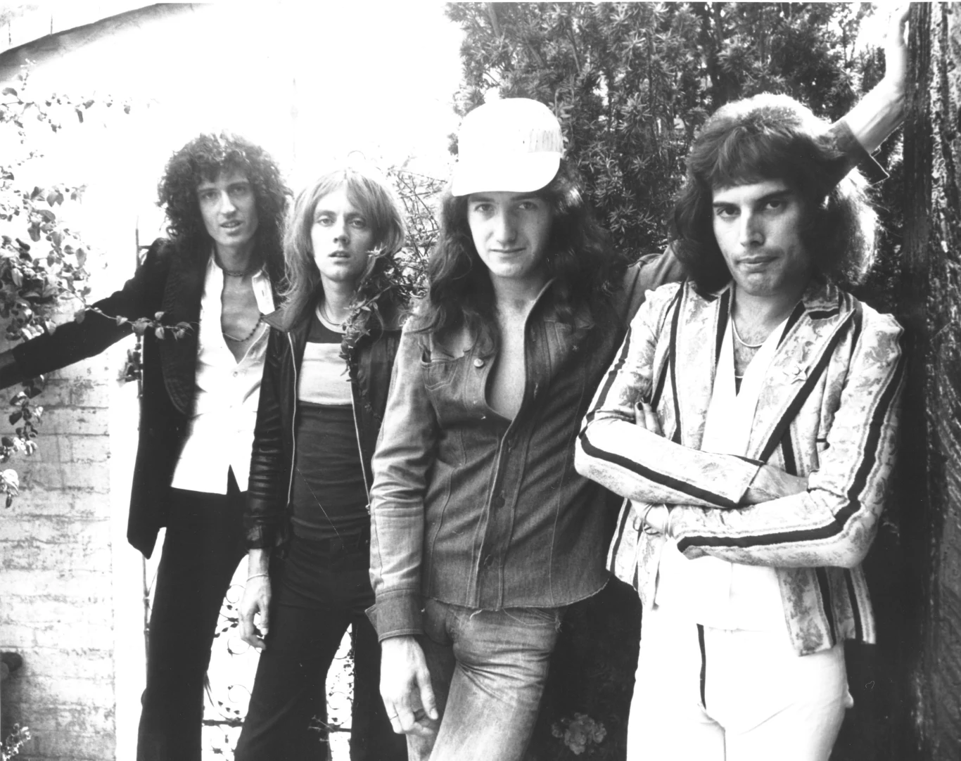 Queen w połowie lat 70. - od lewej: Brian May, Roger Taylor, John Deacon i Freddie Mercury Członkowie zespołu Queen w połowie lat 70., stojący razem, ubrani w różne stylizacje, zdjęcie czarno-białe.