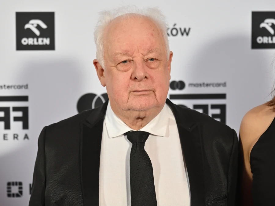 starszy mężczyzna w eleganckim czarnym garniturze i białej koszuli z czarnym krawatem stoi na tle białej ścianki z logotypami sponsorów oraz wydarzenia