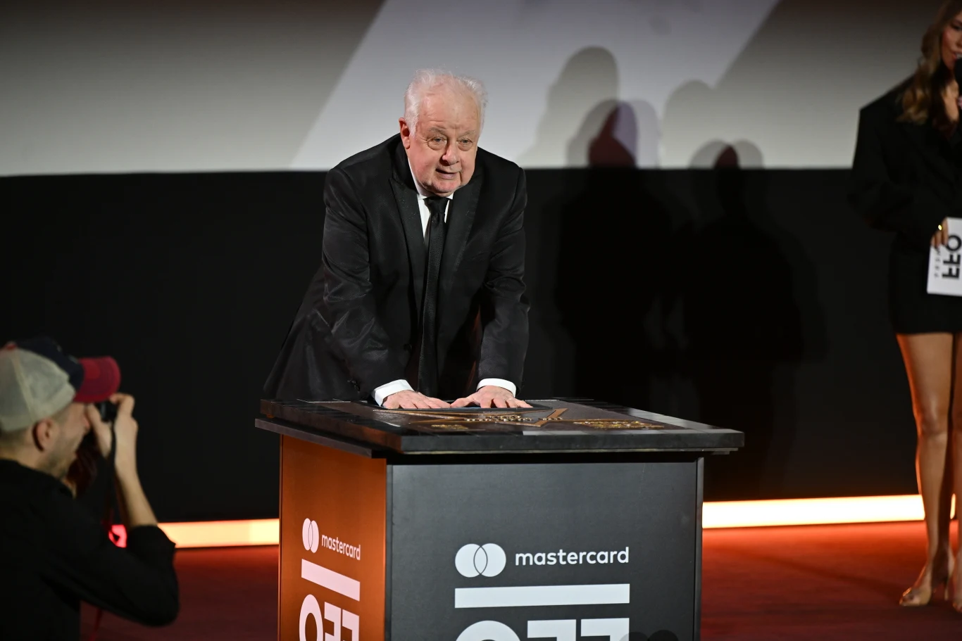 Jim Sheridan podczas gali otwarcia 19. edycji festiwalu Mastercard OFF CAMERA Starszy mężczyzna w garniturze pochyla się nad czarnym podium z logo Mastercard podczas oficjalnego wydarzenia, wokół niego widoczni są fotograf i inni uczestnicy uroczystości w eleganckich strojach.