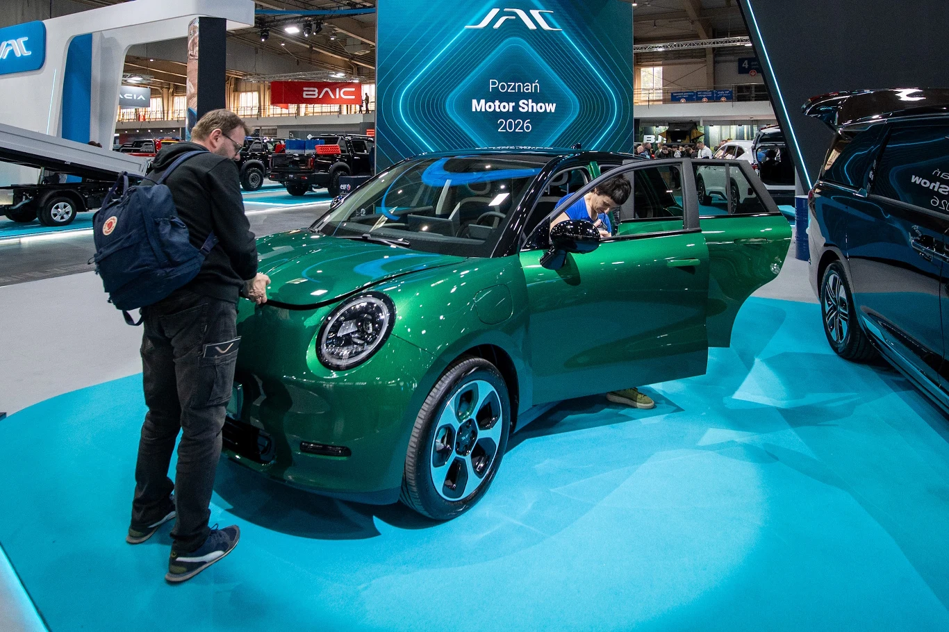 JAC E30X zielony samochód elektryczny prezentowany na stoisku targowym, z otwartymi drzwiami i maską, oglądany przez dwóch mężczyzn zainteresowanych wnętrzem pojazdu, w tle tablica informacyjna z napisem 'Poznań Motor Show 2026'