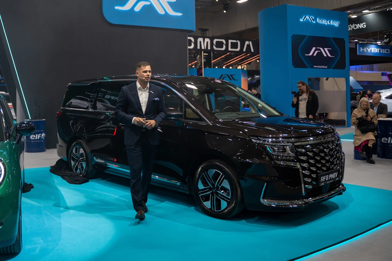 JAC RF8 czarny, nowoczesny minivan marki JAC prezentowany na targach motoryzacyjnych, elegancko ubrany mężczyzna stoi obok pojazdu, w tle widoczne stoiska innych marek oraz kilku zwiedzających