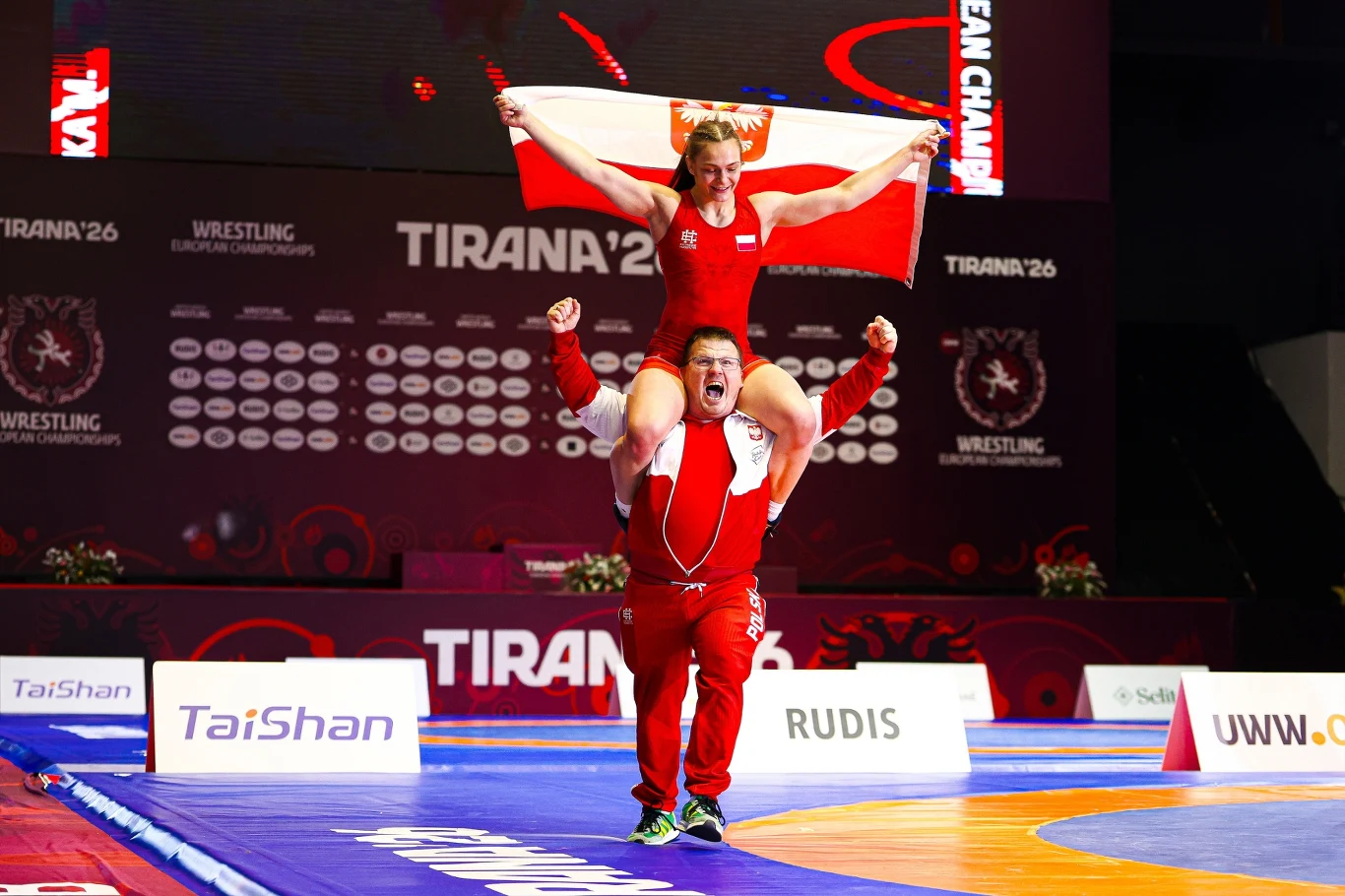 Magdalena Głodek-Liszewska Zawodniczka ubrana w czerwony strój sportowy siedzi na barkach trenera, trzymając nad głową biało-czerwoną flagę, oboje wyrażają radość ze zwycięstwa na tle hali sportowej z napisem 'Tirana '26' i logotypami mistrzostw Europy w zapasach.