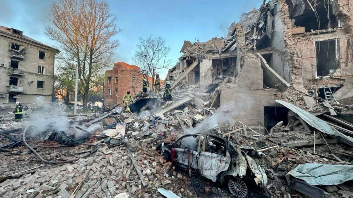 Atak Rosji na Dniepr ruiny zniszczonego budynku, wokół których leżą gruzy i szczątki, spalony samochód w centralnej części obrazu oraz strażacy w oddali prowadzący akcję ratunkową