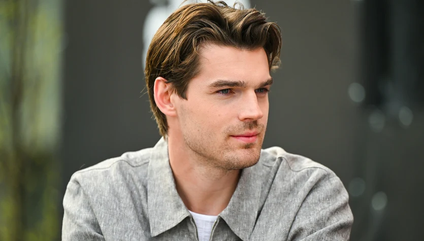 Młody mężczyzna o jasnej karnacji i brązowych, zaczesanych do tyłu włosach w jasnoszarej koszuli, patrzący w bok z lekkim półuśmiechem na tle rozmytego, ciemnego tła.