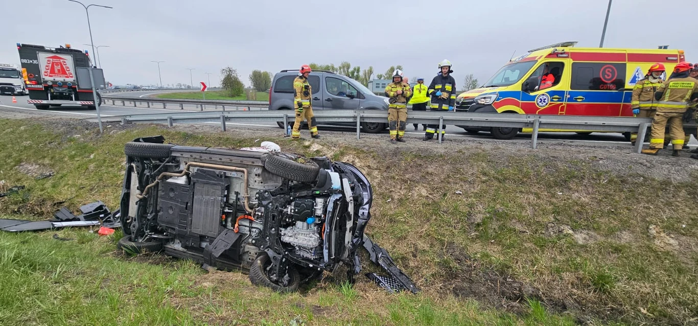 samochód osobowy przewrócony na bok w rowie przy drodze, wokół miejsce wypadku zabezpieczają strażacy, a w pobliżu znajduje się karetka pogotowia oraz samochód strażacki