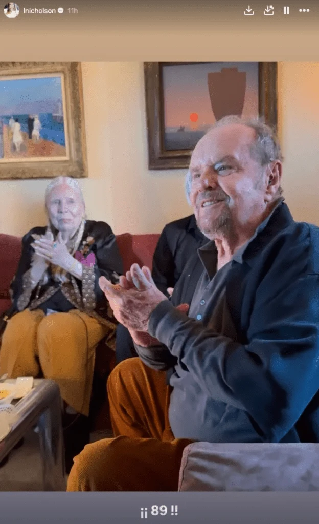 Jack Nicholson w towarzystwie Joni Mitchell Uśmiechnięty, starszy mężczyzna siedzi na kanapie i klaszcze, obok niego kobieta w zbliżonym wieku również klaszcze, w tle widoczne są dwa obrazy na ścianie oraz niewielki stolik z papierami i kubkiem.
