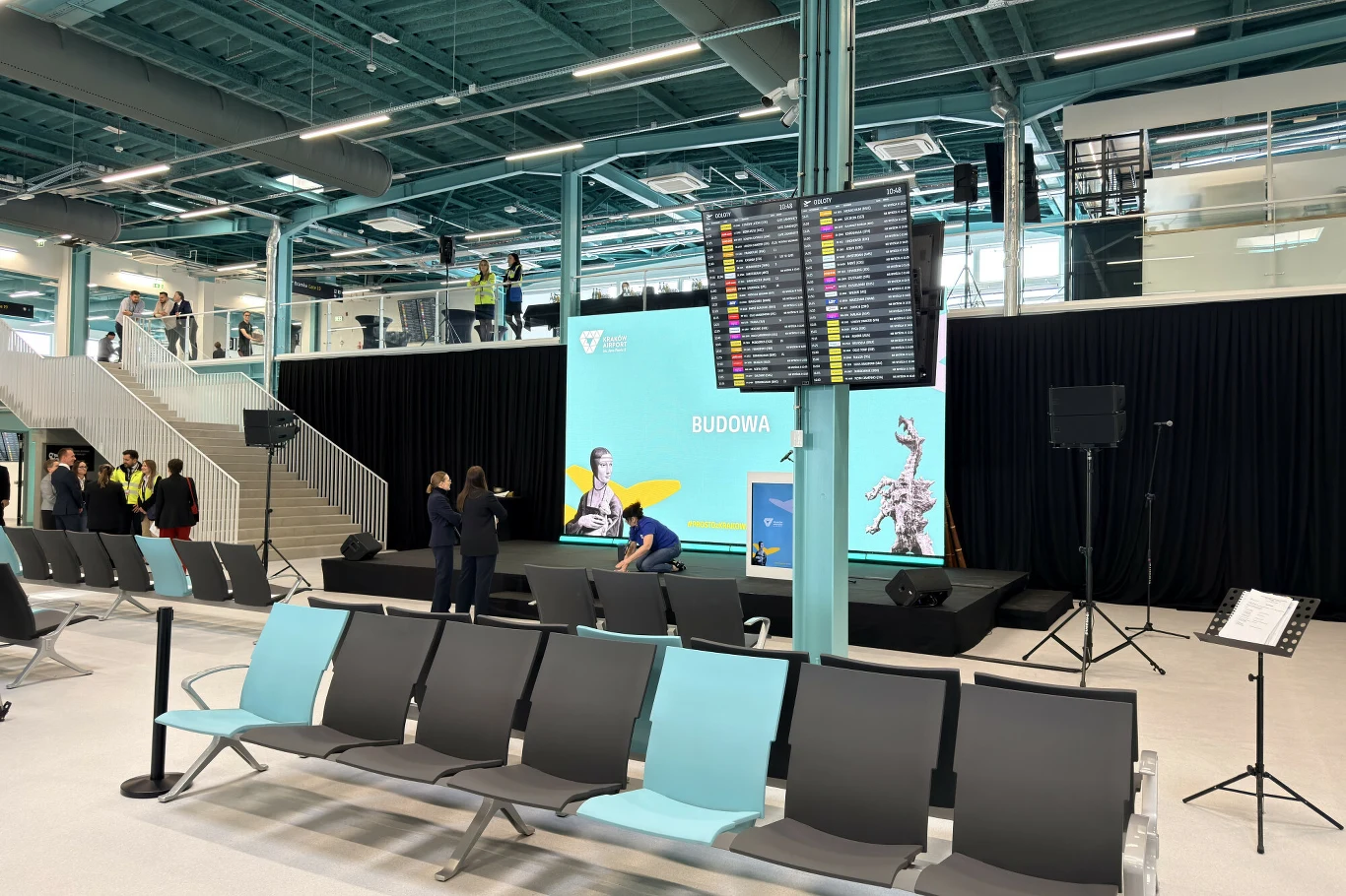 Z magazynu w terminal. Tak zmieniło się lotnisko w Krakowie Wnętrze nowego terminala w Krakowie Balicach. Rzędy foteli, ekran informacyjny, scena z napisem 'Budowa'.