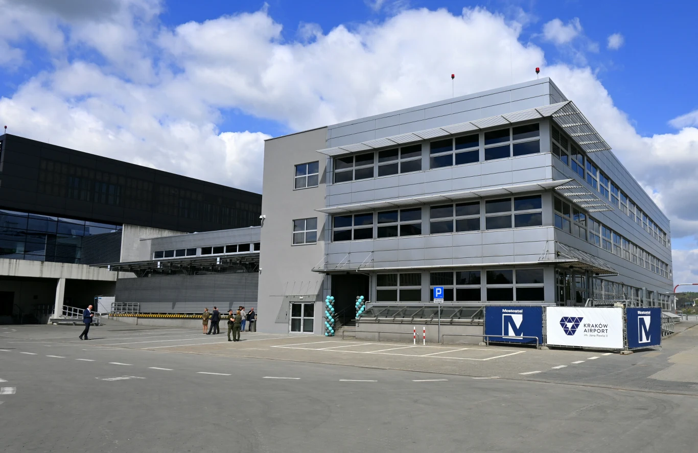 Międzynarodowy Port Lotniczy Kraków-Balice. Nowy Terminal Wspierający - dawny budynek Cargo Nowoczesny budynek terminala lotniska z przeszklonymi fasadami, dużymi oknami i srebrnymi akcentami, przed którym stoi kilka osób oraz oznaczone są wejścia i informacje związane z lotniskiem Kraków Airport.
