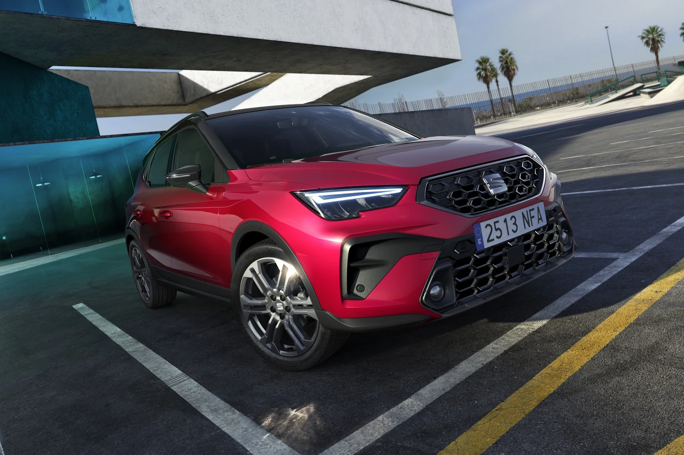 Seat Arona ma nowe kolory i niższe ceny Czerwony Seat Arona na betonowym parkingu, w tle nowoczesny budynek i palmy.