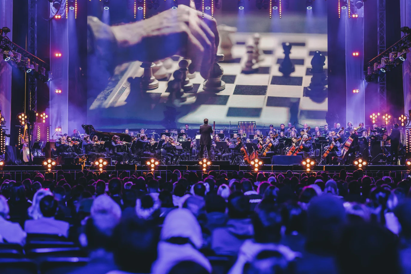 orkiestra symfoniczna grająca na scenie przed dużą publicznością, nad sceną wyświetlony jest ogromny ekran pokazujący zbliżenie dłoni przesuwającej figurę szachową na szachownicy