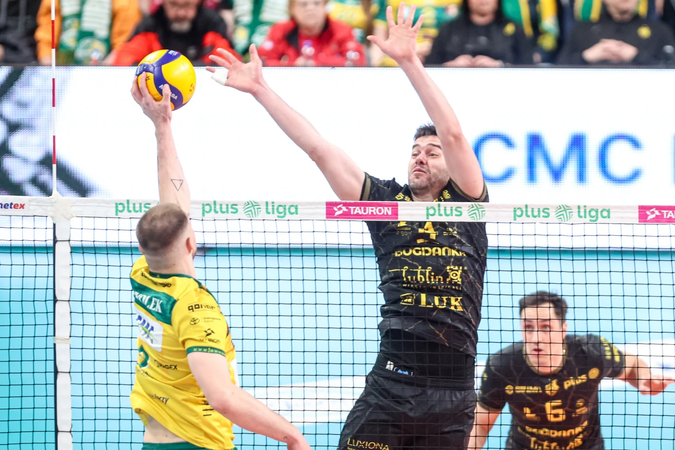 zawodnik w żółto-zielonym stroju wykonuje atak siatkarski, natomiast przeciwnik w czarnej koszulce próbuje go zablokować przy siatce; w tle widoczni są kibice uważnie obserwujący akcję