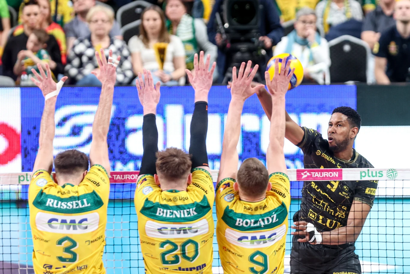 zawodnik w czarnym stroju atakuje piłkę siatkową mocnym uderzeniem, próbując przebić się przez potrójny blok trzech siatkarzy w żółto-zielonych koszulkach z nazwiskami Russell, Bieniek i Bołądź, w tle widownia skupiona na przebiegu meczu