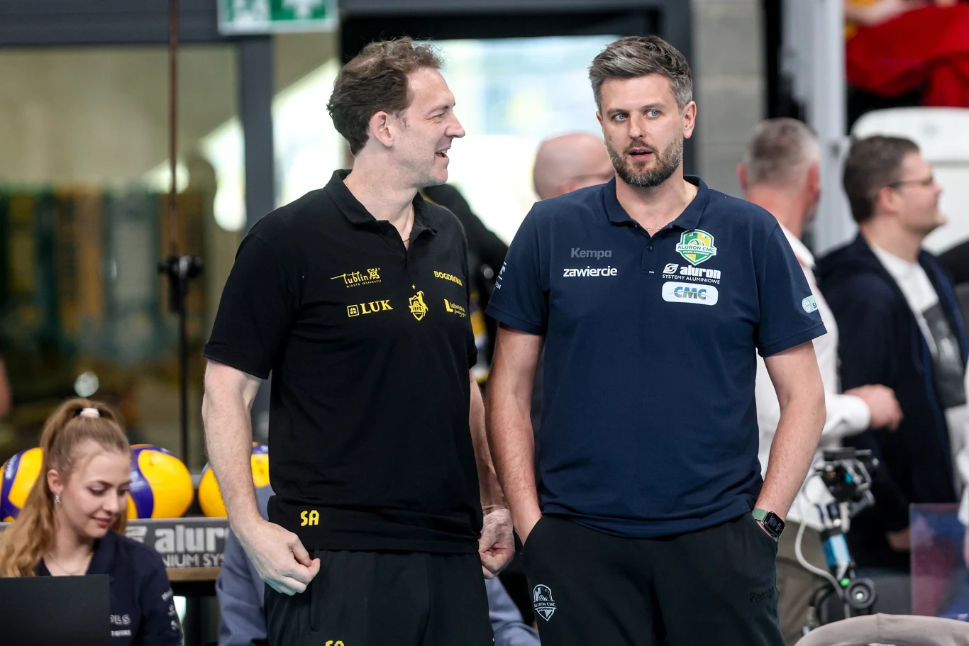 Dwóch mężczyzn w sportowych koszulkach drużyn siatkarskich rozmawia ze sobą podczas meczu, w tle znajdują się inni członkowie sztabu i zawodnicy oraz siatkarskie piłki.