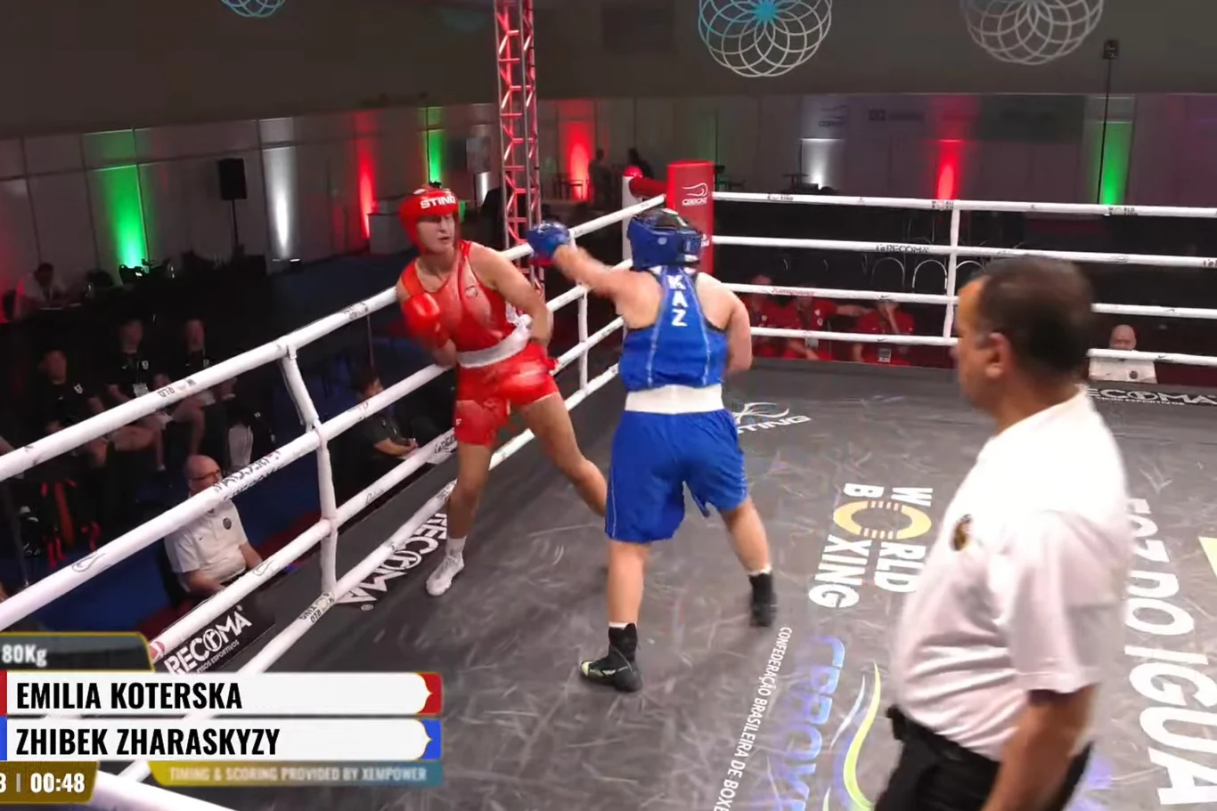 Emilia Koterska (w czerwonym stroju) w walce o finał PŚ zawodniczka w niebieskim stroju zadaje cios przeciwniczce uwięzionej w rogu ringu, sędzia uważnie obserwuje przebieg walki
